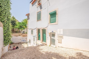 A narrow cobblestone street with a white building featuring green shutters and a visible door. - GuestReady - The George (Lisbon)