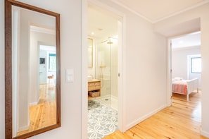 A hotel room with a wooden floor, a bathroom with a glass shower, and a bed with a striped blanket. - GuestReady - The George (Lisbon)
