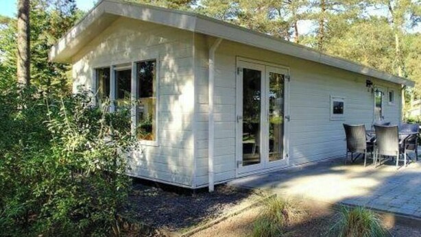 A single-story, light-colored cabin with a porch, surrounded by trees. - Unique Nouveau 4 (Otterlo)