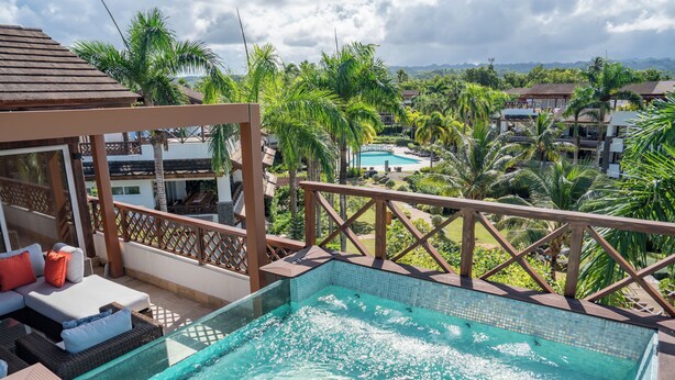 A rooftop pool with a wooden deck, lounge area with cushions, and a view of a resort with palm trees and buildings in the background. - Unique Penthouse with Huge Private Rooftop & Jacuzzi - Beach & Restaurant Nearby (El Limón)