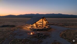 A lodge with a large central room and a deck, surrounded by a dry, open landscape with mountains in the background.
