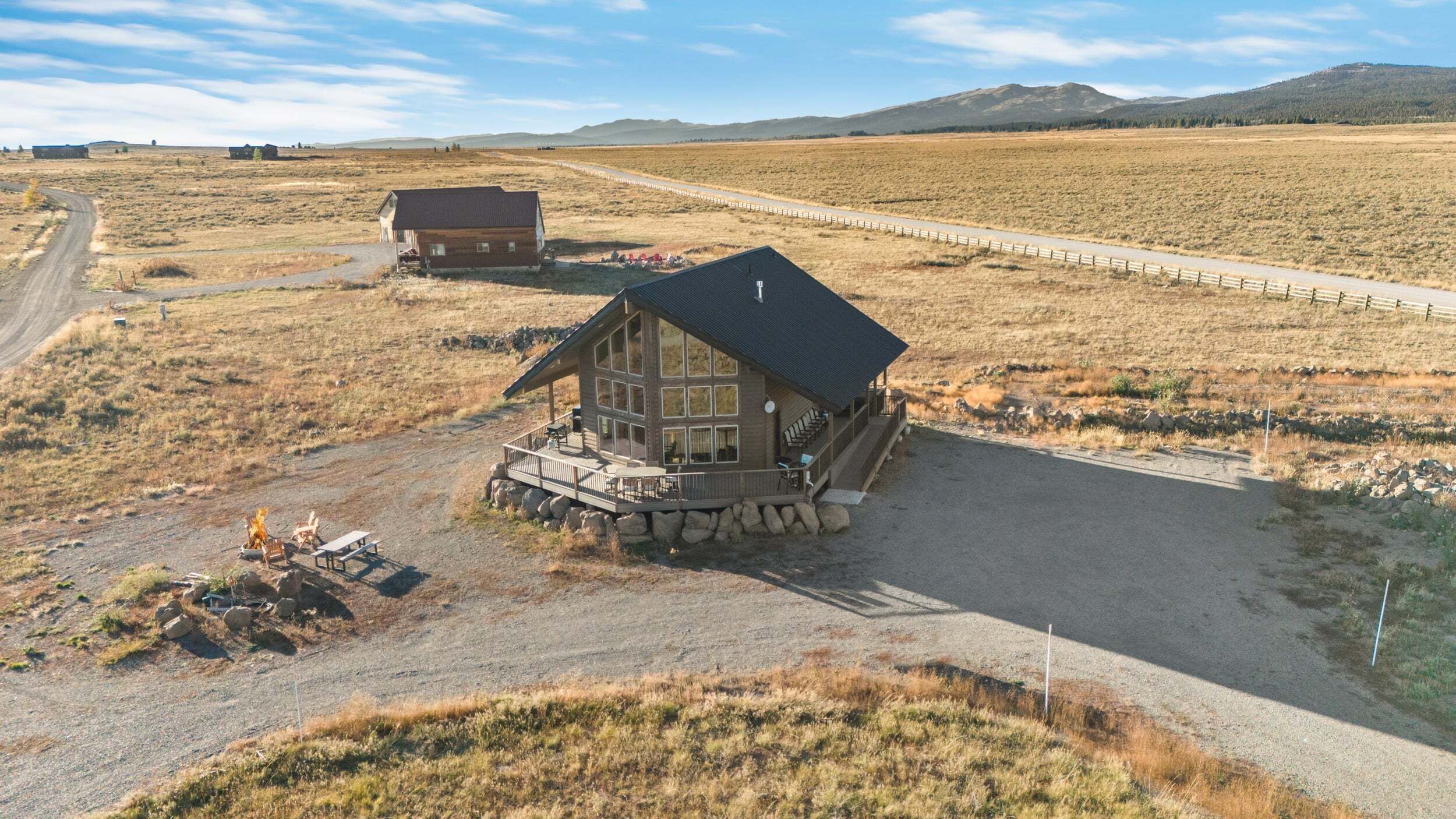 A log cabin with a porch and a deck, surrounded by a gravel driveway and a dirt path.