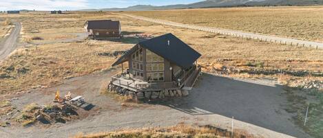 A log cabin with a porch and a deck, surrounded by a gravel driveway and a dirt path.