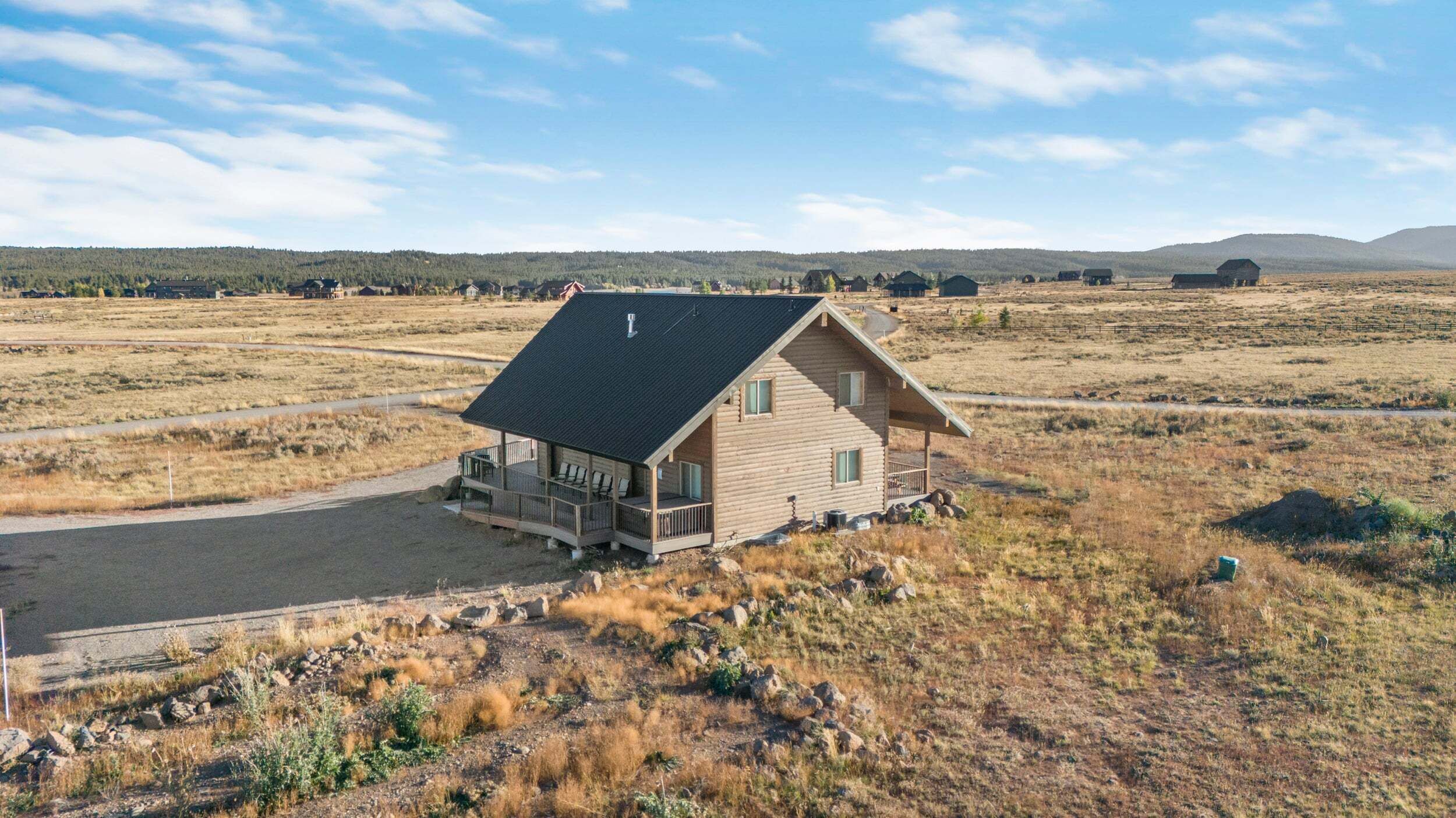 A log cabin with a porch, surrounded by a dry, grassy landscape with distant hills.