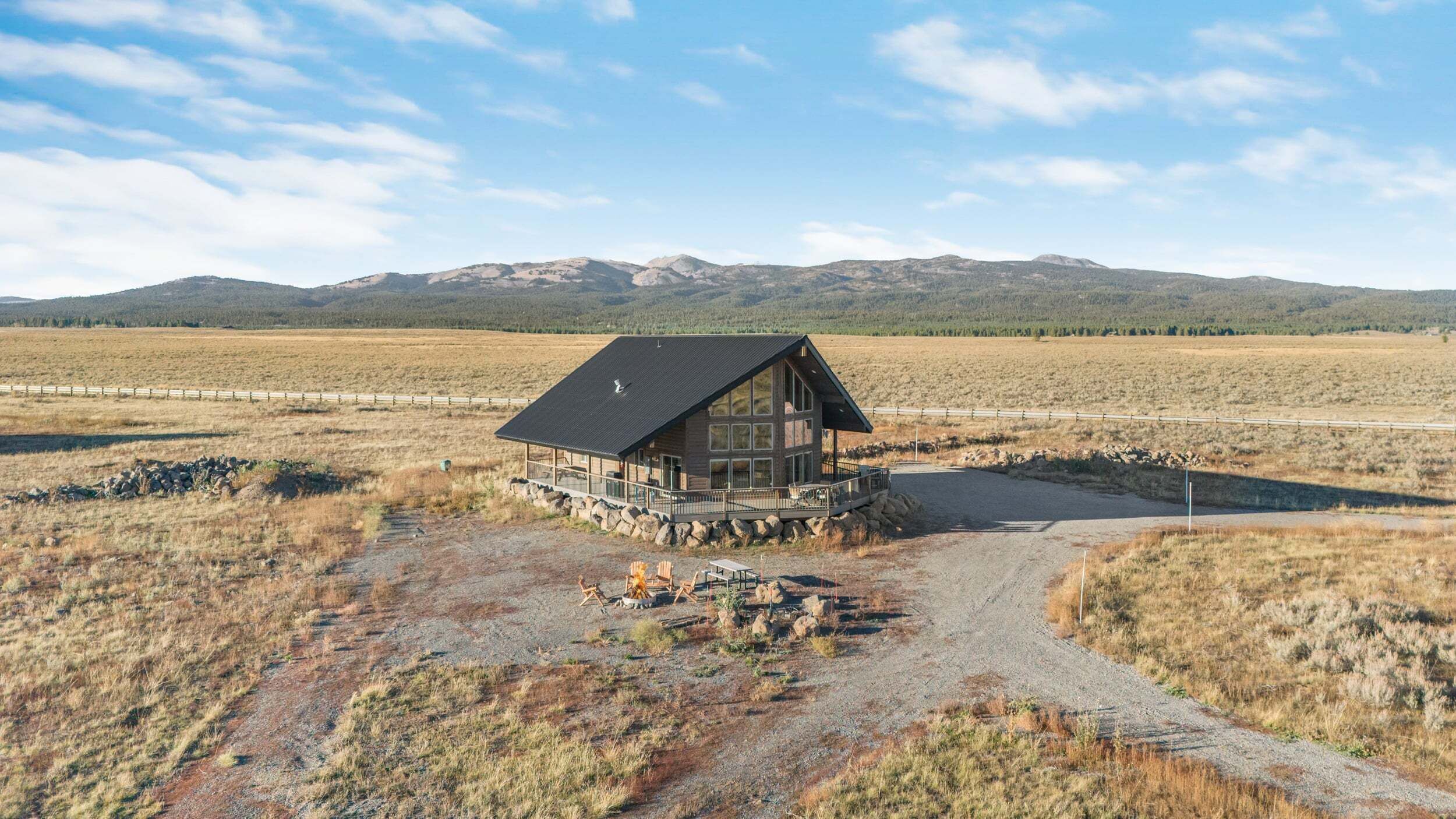 En hytte med stor terrasse, omgivet af en grussti og et træhegn, med bjerge i baggrunden.