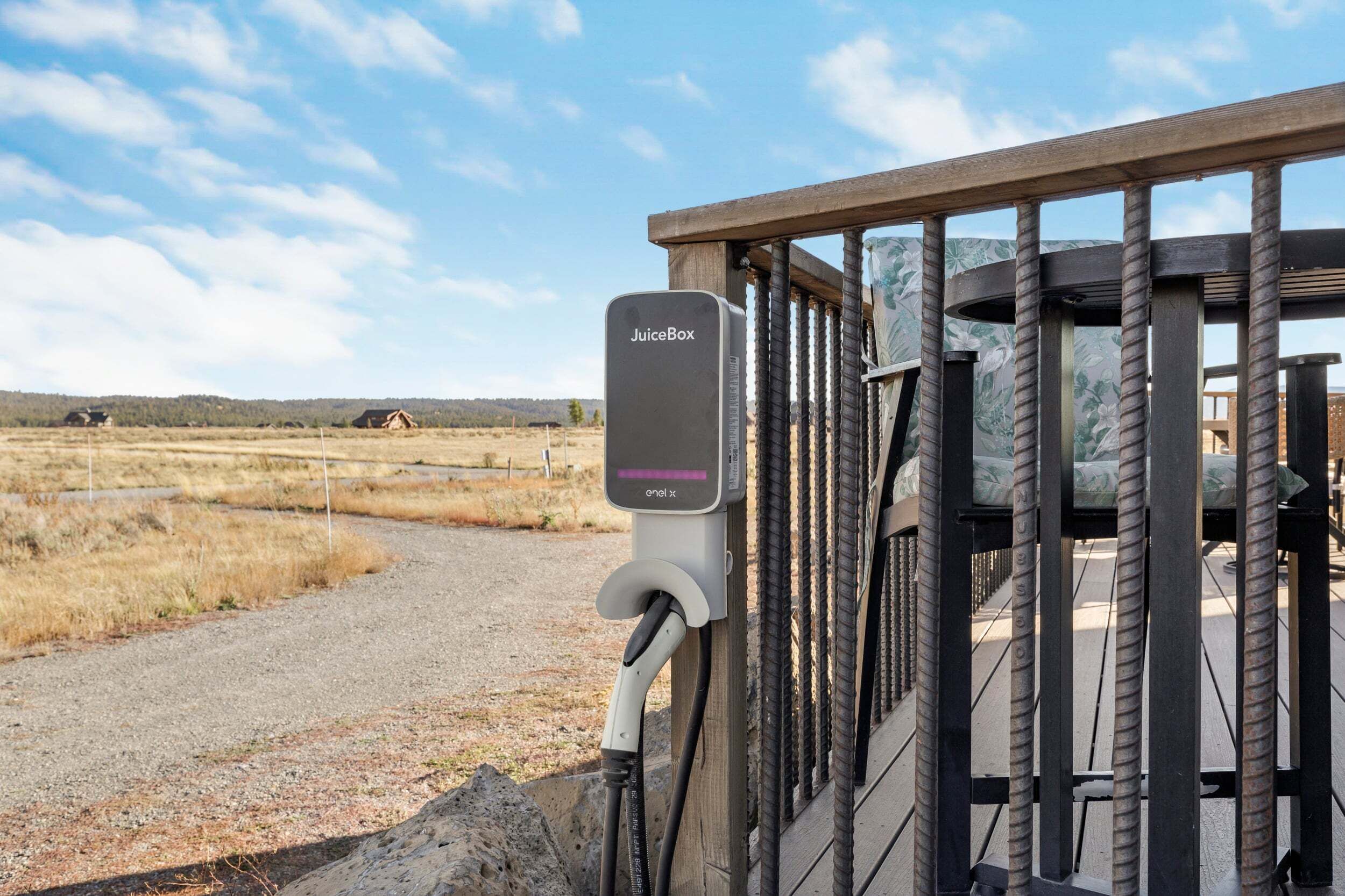 A JuiceBox charging station attached to a wooden fence with a gravel path and distant buildings.