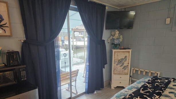 A room with a bed, a TV mounted on the wall, a refrigerator, a microwave, a bench, and a view through a glass door.