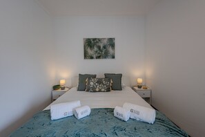 Ein ordentlich bezogenes Bett mit blauen, gemusterten Bettdecken, zwei Kissen, zwei Nachttischlampen und einem Wandbild mit tropischem Motiv.