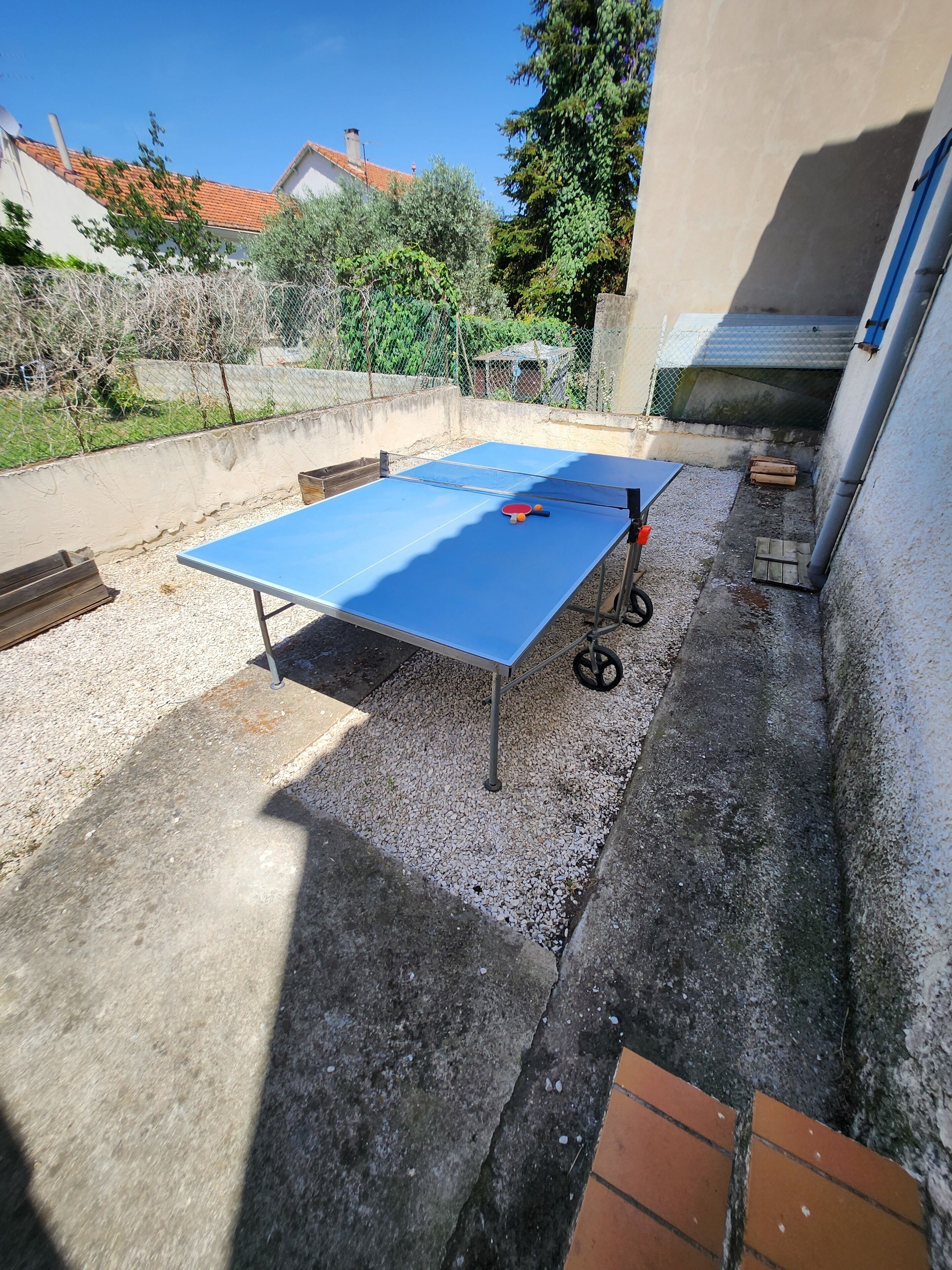 An outdoor ping pong table with a net, surrounded by gravel and a concrete floor, with a building and trees in the background.