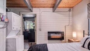 A bedroom with a bed, a bedside table with a lamp, a wall-mounted fireplace, and a washing machine. - Shotgun Chalet (Island Park)