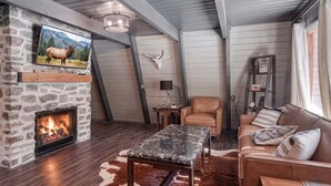 A cozy living room with a stone fireplace, a mounted TV, a leather sofa, and a cowhide rug. - Shotgun Chalet (Island Park)