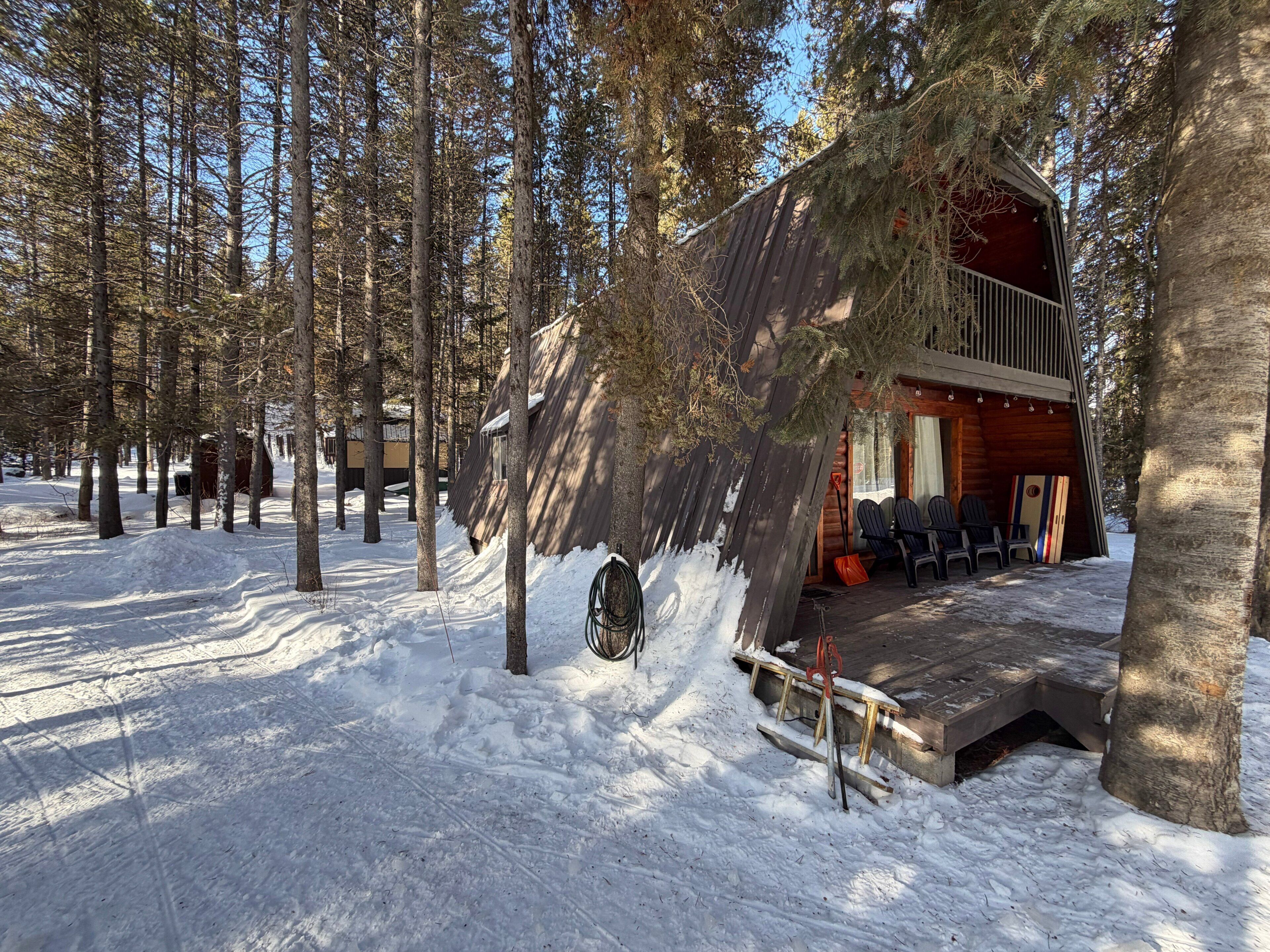 Una cabaña en un bosque nevado con una terraza y sillas afuera.