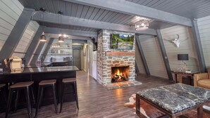 A loft-style room with a stone fireplace, wooden beams, a kitchen area, and a bar with stools. - Shotgun Chalet (Island Park)