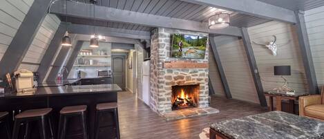 A loft-style room with a stone fireplace, wooden beams, a kitchen area, and a bar with stools.