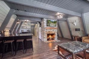 A loft-style room with a stone fireplace, wooden beams, a kitchen area, and a bar with stools.