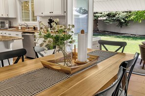 A modern kitchen with white cabinets, a wooden dining table with a placemat, a vase with flowers, and a paper towel holder.