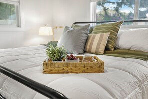 A neatly made bed with a white comforter, a wicker tray with a small plant and fruit, and pillows in shades of green and gray.