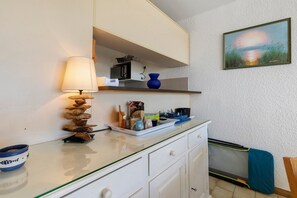Una cocina con mostrador blanco, lámpara colgante, un florero azul y un cuadro en la pared.