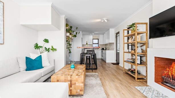 Een moderne woonkamer met een bank, salontafel en open haard. Achtergrond toont een keuken.