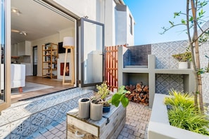 Ein modernes Haus mit Terrasse, einer hölzernen Bank, Blumentöpfen und einem sichtbaren Küchenbereich.