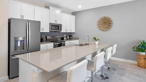 A modern kitchen with white cabinets, stainless steel appliances, a central island with bar stools, and a decorative wall piece. - Go!#32 CHAMPIONS GATE (Kissimmee)