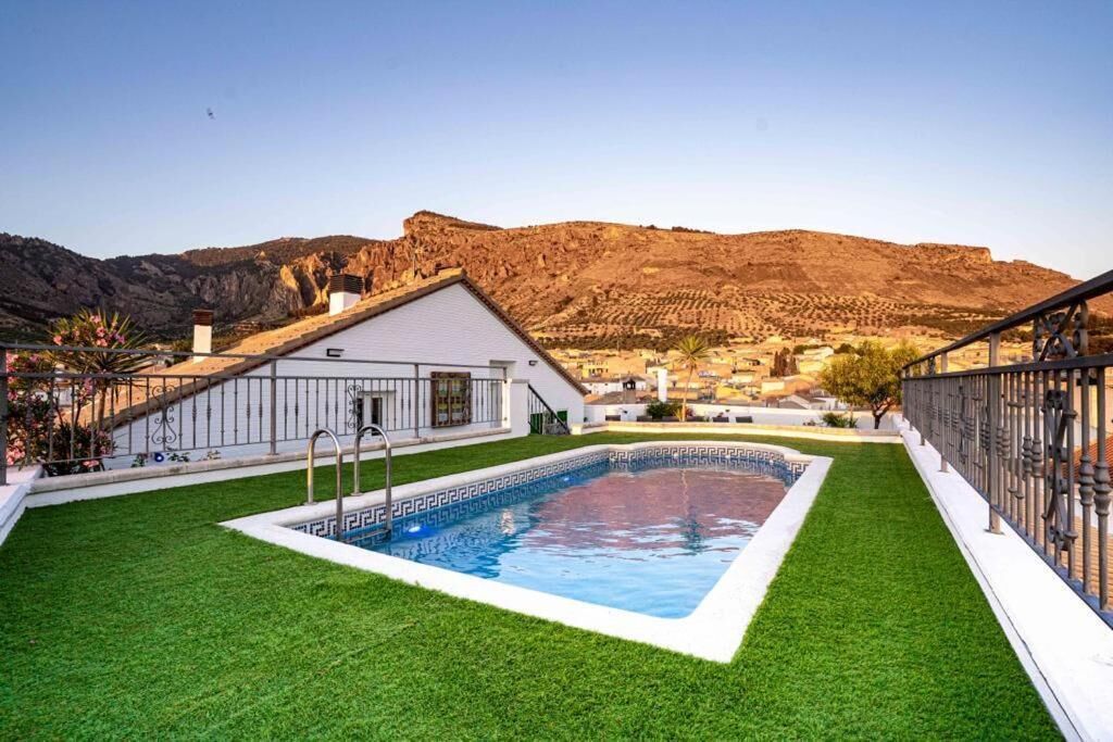 A swimming pool with a white border, surrounded by green artificial grass, set against a mountainous backdrop.