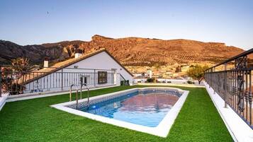 A swimming pool with a white border, surrounded by green artificial grass, set against a mountainous backdrop.