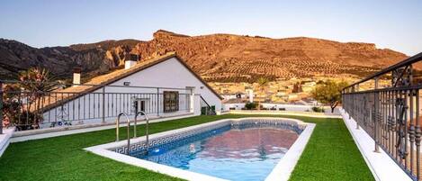 Una piscina con borde blanco, rodeada de césped artificial verde, con montañas al fondo.