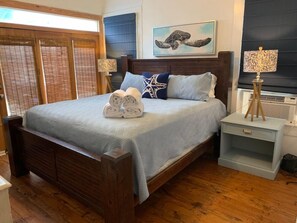 A bedroom with a wooden bed, a nightstand with a lamp, a window with blinds, and a wall-mounted air conditioner. - 2423 Q5 Near the Pier (Galveston)