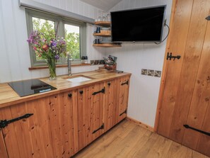 A kitchen with wooden cabinets, a built-in stove, a window with a flower vase, and a mounted TV. - Bramleys (Watchet)