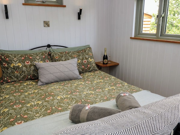 A bed with floral bedding, a bedside table with a bottle and glasses, and a window with a view of greenery. - Bramleys (Watchet)
