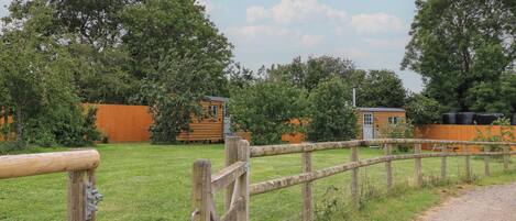 Entorno rural con valla de madera, camino de grava y dos cabañas de madera con construcciones adosadas.