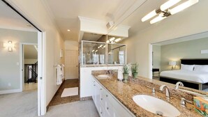Un baño moderno con encimera de granito, dos lavamanos, un espejo grande y una habitación separada visible a través de una puerta abierta.