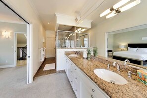 Une salle de bain moderne avec un comptoir en granit, deux lavabos, un grand miroir et une chambre à coucher séparée, visible à travers une porte ouverte.