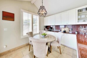 A dining area with a round table, chairs, a fruit bowl, and a kitchen with white cabinets and a marble countertop.