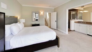 Un dormitorio con cama, mesita de noche, lámpara, espejo, puerta y baño con lavabo y espejo.
