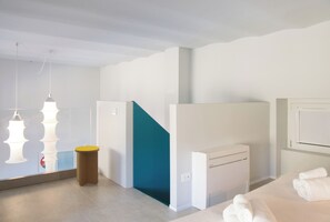 Une chambre d’hôtel moderne, équipée d’un lit, d’un radiateur, d’un petit tabouret et d’un miroir qui reflète une partie de la pièce.