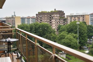 Ein Balkon mit Tisch und Stühlen, der auf einen dicht bebauten Stadtbereich mit mehrstöckigen Gebäuden und üppigen Grünflächen blickt.