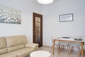 A room with a beige sofa, a dining table with four chairs, a white round table, and a door with a glass panel. - Bright Nest Bande Nere – Milano Apartment (Milano)