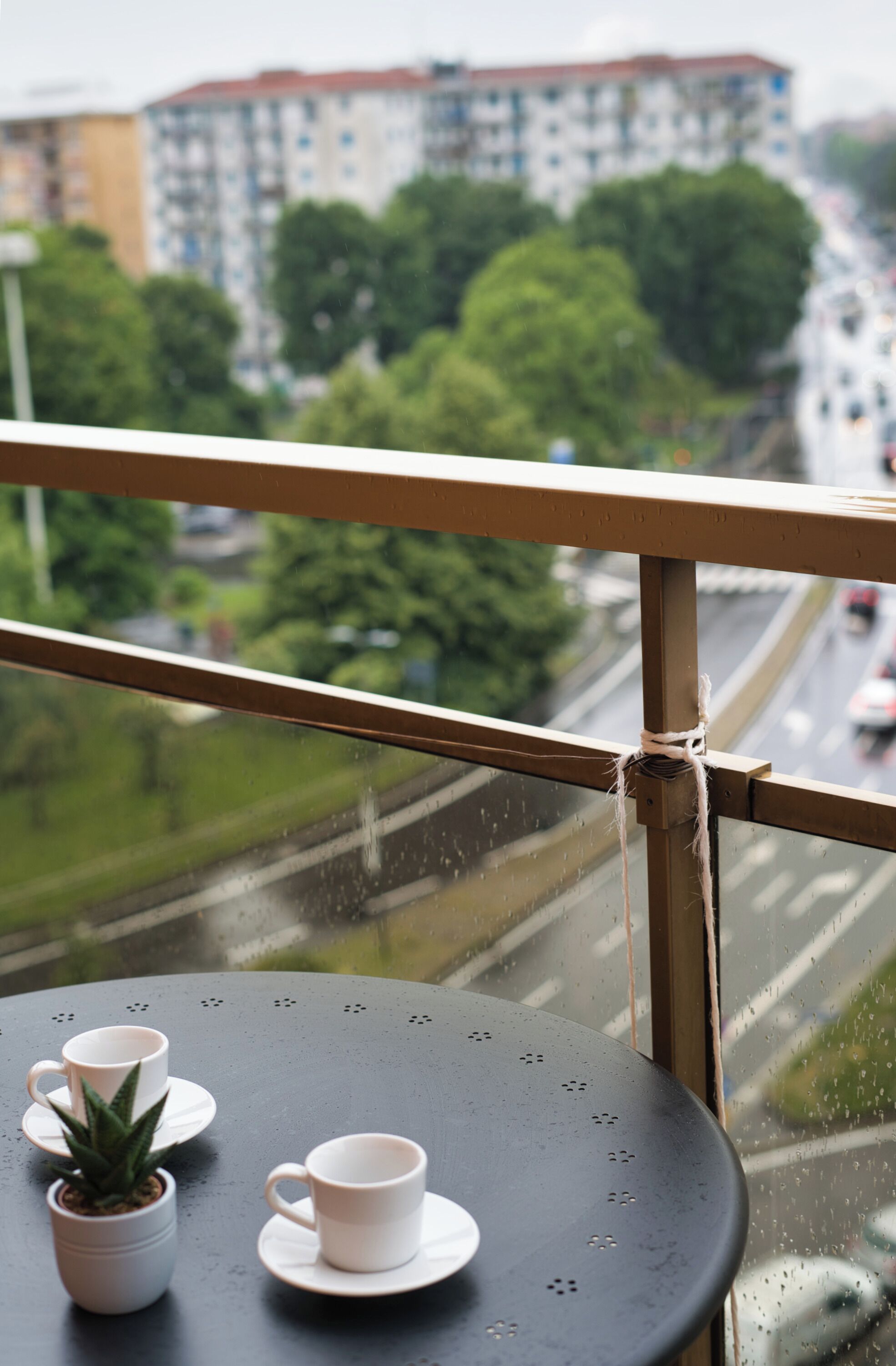 A balcony with a view of a cityscape, a table with two cups and a small plant, and a rope tied to the railing.
