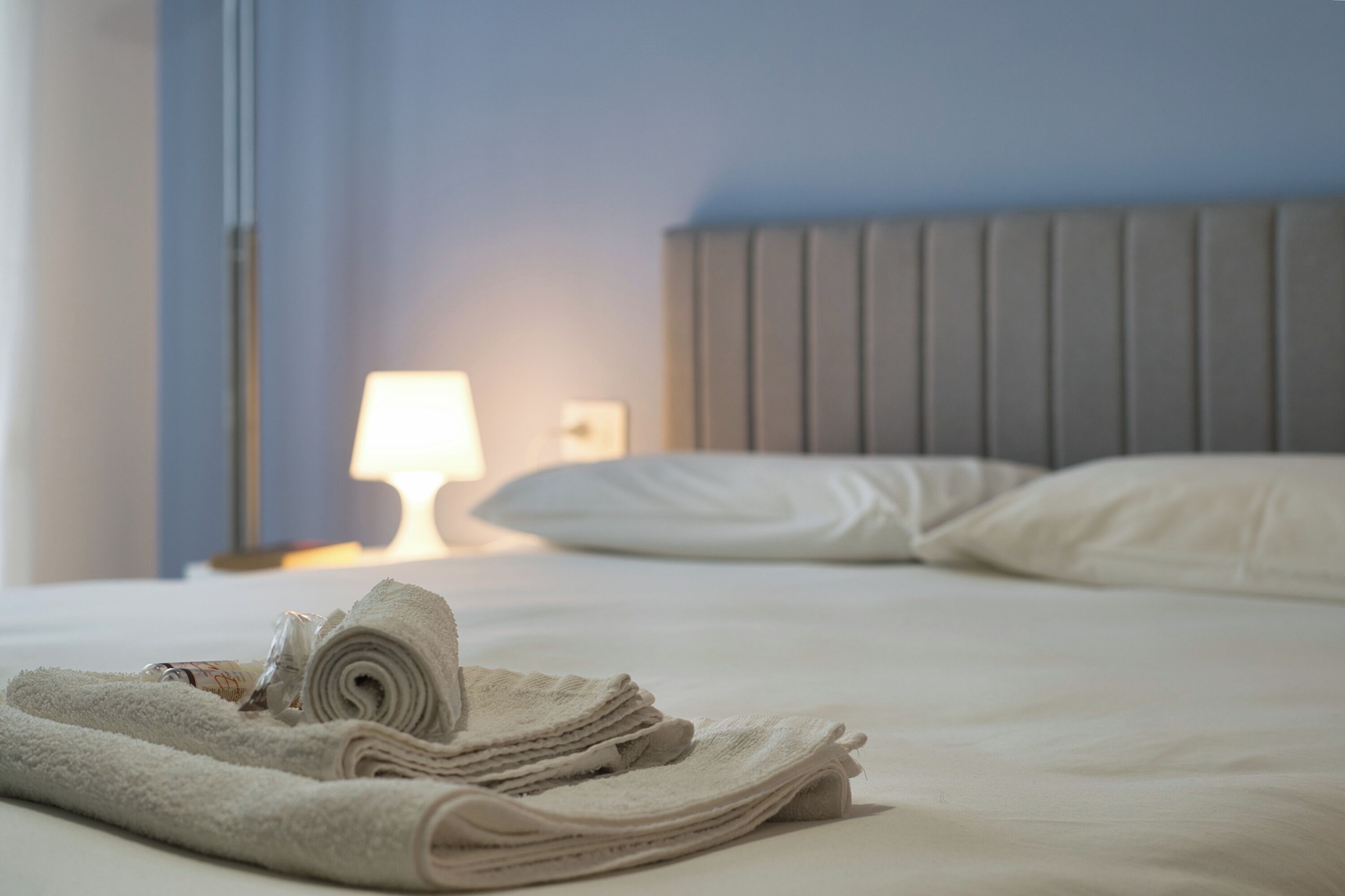 Ein ordentlich bezogenes Bett mit aufgerollten Handtüchern, einer Nachttischlampe und einem Kopfteil.