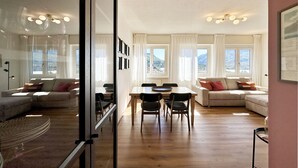 A spacious living area with a dining table, multiple sofas, and large windows with curtains.