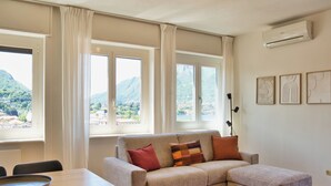 A living room with a sofa, a dining table, and chairs. There are large windows with curtains, an air conditioning unit, and framed artwork on the wall.