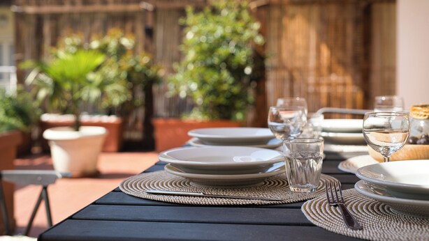 Mesa de comedor al aire libre dispuesta con platos, vasos y cubiertos, rodeada de plantas en macetas y una valla de madera.