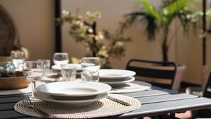 Una mesa de comedor dispuesta con platos, vasos y cubiertos, con una silla y una planta en maceta al fondo.