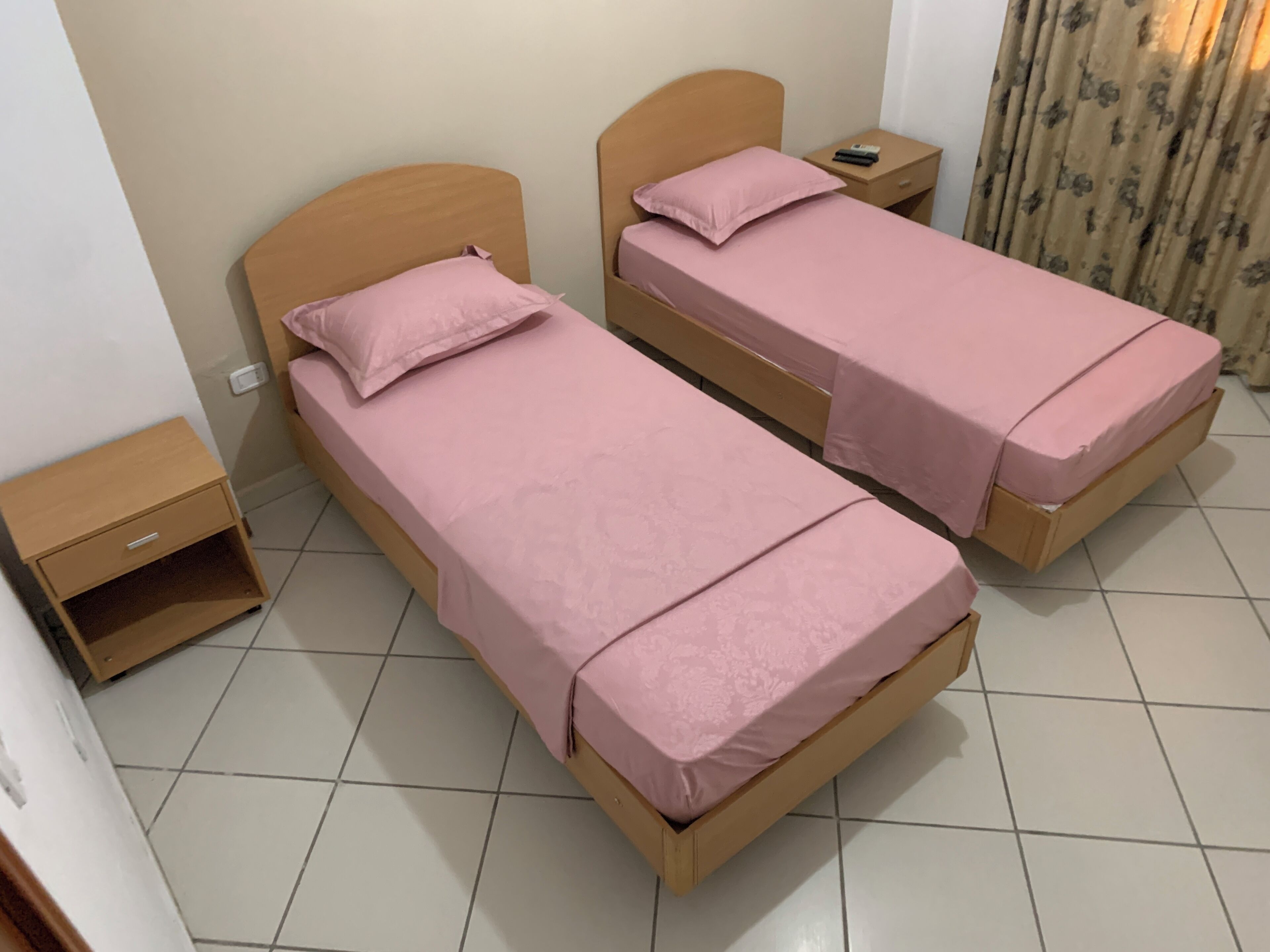 Two single beds with wooden headboards, a small bedside table with a remote control, and a curtain on the right side.