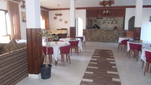 Een restaurantgedeelte in een hotel met ronde tafels, rode stoelen, een kamerbreed tapijt en een bar op de achtergrond.