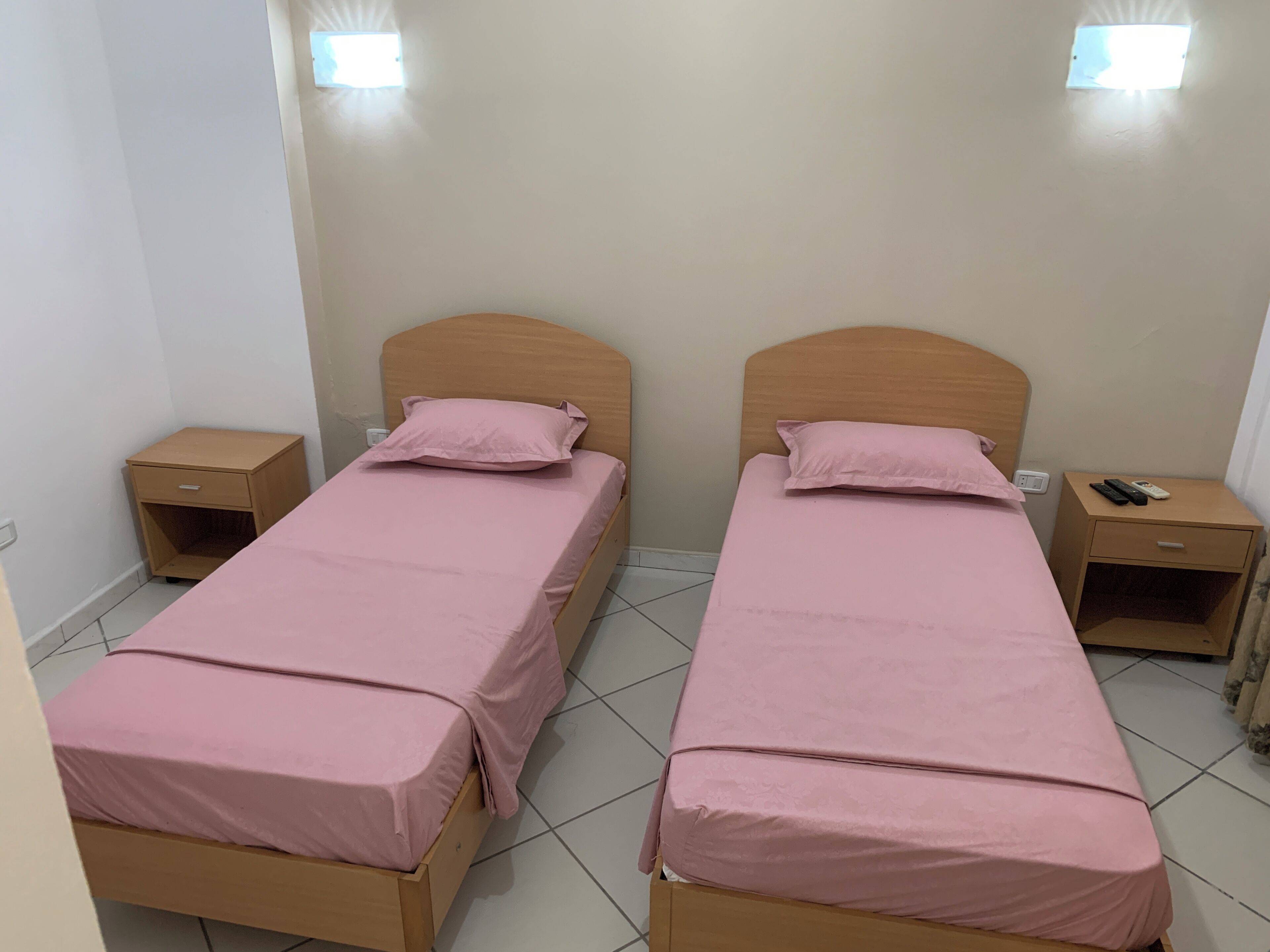 Quarto duplo com duas camas, cada uma com cabeceira de madeira, mesa de cabeceira e luminária na parede.