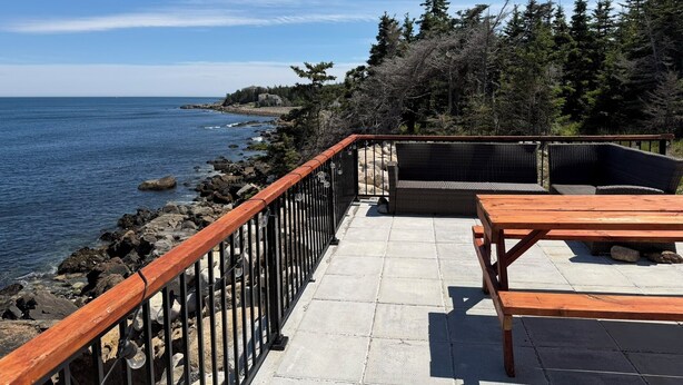 A coastal patio with a wooden table and chairs, a stone pathway, and a view of the sea and trees.