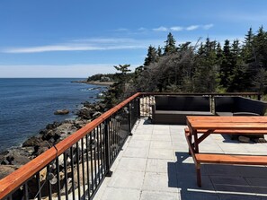 Un patio frente al mar con mesa y sillas de madera, un sendero de piedra y vistas al mar y árboles.